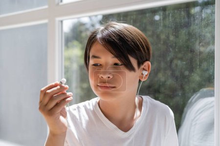 Asiatisches Kind schaut auf kabelgebundenen Kopfhörer in der Nähe des Fensters zu Hause 
