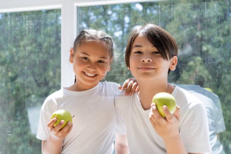Foto de Sonrientes hermanos asiáticos sosteniendo manzanas frescas cerca de ventana en casa - Imagen libre de derechos
