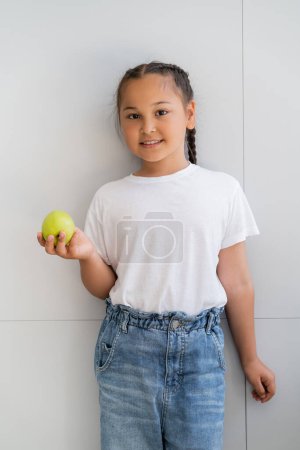 Lächelndes asiatisches Mädchen mit reifem Apfel und Blick auf Kamera in der Nähe der Hauswand 