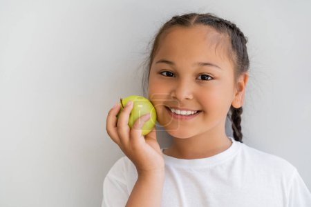 Portrait of smiling asian girl holding fresh apple and looking at camera near wall 