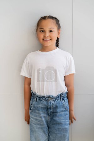 Cheerful asian child in casual clothes standing near wall at home 