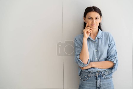 Mujer morena en camisa y jeans mirando a la cámara cerca de la pared gris 
