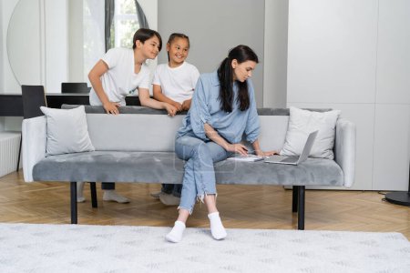 Positive asian kids looking at mother working on laptop on sofa at home 