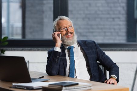Positive bearded businessman in eyeglasses talking on smartphone in office 