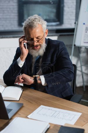 Photo for Middle aged businessman talking on smartphone and looking at wristwatch in office - Royalty Free Image