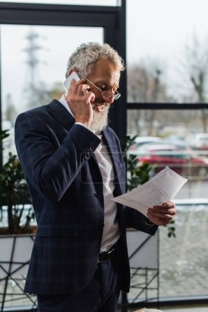 Smiling mature businessman talking on smartphone and holding blueprint in office 