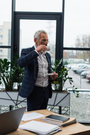 Geschäftsmann mittleren Alters trinkt Kaffee in der Nähe von Laptop und Papieren im Büro 