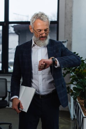 Mature businessman in suit looking at wristwatch and holding notebook in office 