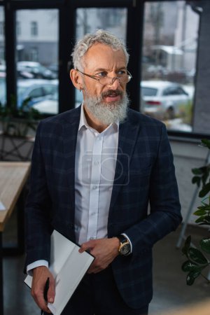 Photo for Bearded mature businessman in suit holding notebook in office - Royalty Free Image