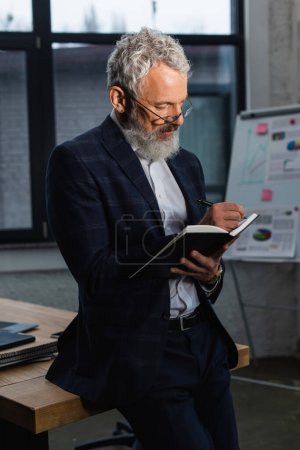 Reifer Geschäftsmann im Anzug schreibt im Büro auf Notizbuch 