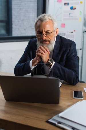 Konzentrierter reifer Geschäftsmann blickt im Büro auf Laptop in der Nähe von Zeitungen und Smartphone 