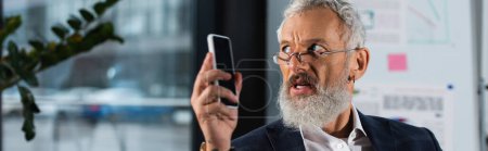 Photo for Middle aged businessman in eyeglasses holding smartphone and looking away in office, banner - Royalty Free Image