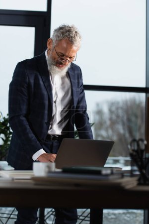 Mature manager in suit standing near laptop on table in office in evening 