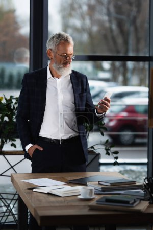 Reifer Geschäftsmann im Anzug mit Smartphone in der Nähe von Notebooks und Kaffee im Büro 