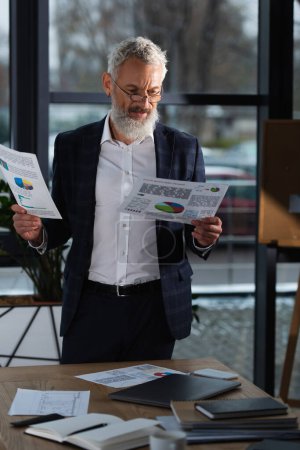 Foto de Hombre de negocios de mediana edad mirando gráficos cerca de mesa de trabajo en la oficina - Imagen libre de derechos