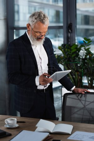 Grauhaariger Geschäftsmann im Anzug mit digitalem Tablet in der Nähe von Kaffee und Notizbuch im Büro 