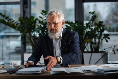 Téléchargez les photos : Homme d'affaires aux cheveux gris en costume prenant des papiers près des appareils et du café au bureau le soir - en image libre de droit