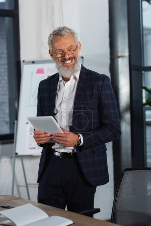 Cheerful grey haired businessman holding digital tablet near notebook on table in office 