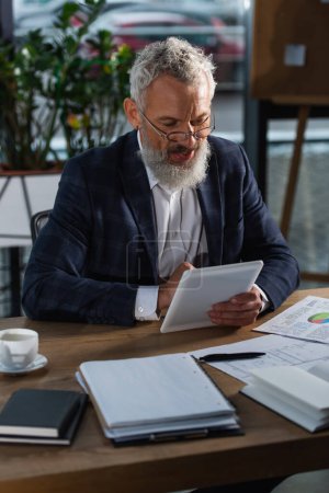 Lächelnder grauhaariger Geschäftsmann mit digitalem Tablet in der Nähe von Dokumenten und Kaffee im Büro 