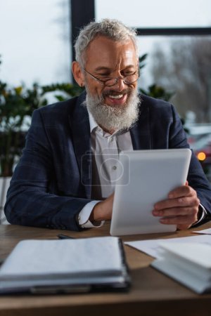 Fröhlicher grauhaariger Geschäftsmann im Anzug mit digitalem Tablet in der Nähe von Zeitungen auf dem Tisch 