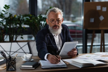 Mature businessman in suit holding digital tablet near documents and coffee in office 