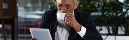 Reifer Geschäftsmann trinkt Kaffee und schaut abends auf digitales Tablet im Büro, Banner 