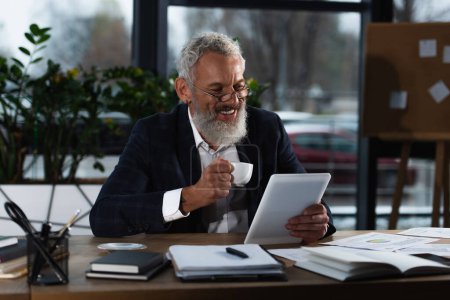 Fröhlich gereifter Geschäftsmann hält Kaffeetasse in der Hand, während er digitales Tablet in der Nähe von Dokumenten im Büro benutzt 