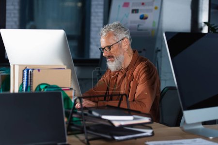 Hombre de negocios maduro en ropa casual y anteojos usando computadora cerca de cuadernos en la oficina 