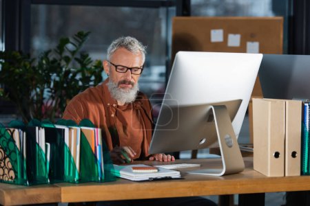 Bearded businessman in eyeglasses sitting near computer and paperwork in office in evening 