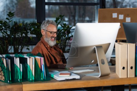 Alegre hombre de negocios de pelo gris que trabaja cerca de la computadora y los papeles en la oficina 