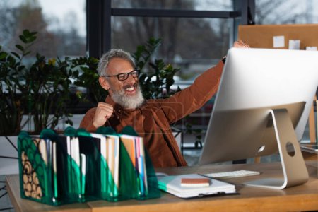 Emocionado hombre de negocios de pelo gris mostrando sí gesto cerca de la computadora y el papeleo en la oficina 