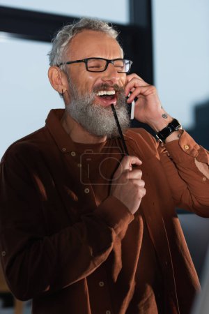Photo for Cheerful bearded businessman in eyeglasses holding pencil while talking on smartphone in office in evening - Royalty Free Image