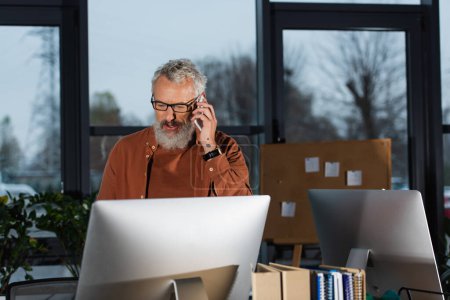 Mature businessman talking on smartphone and looking at computer monitor while working in office in evening 