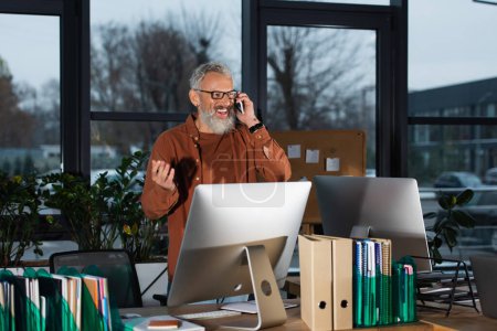 Photo for Positive grey haired manager talking on mobile phone near computers and paperwork in office in evening - Royalty Free Image