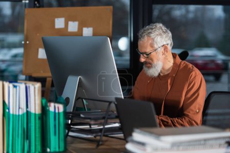 Hombre de negocios maduro en anteojos trabajando cerca de computadoras y documentos borrosos en la oficina 