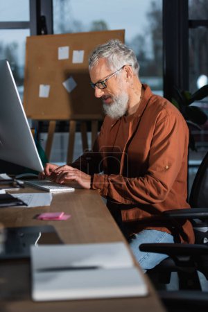 Administrador de pelo gris utilizando la computadora cerca de papeles y cuaderno en la oficina por la noche 