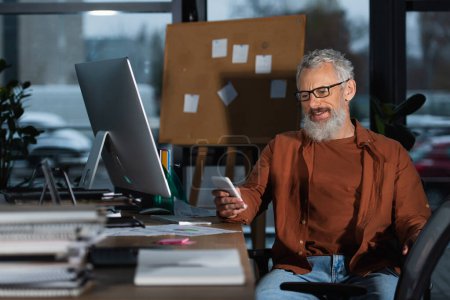 Hombre de negocios de pelo gris sonriente en gafas con teléfono inteligente cerca de la mesa de trabajo por la noche 