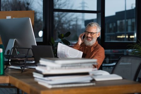 glücklicher Geschäftsmann mittleren Alters, der Dokumente in der Hand hält und in der Nähe von Computern und Notebooks mit dem Smartphone spricht