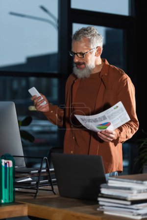 Photo for Smiling bearded businessman holding smartphone and paper with charts near computers in office - Royalty Free Image