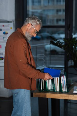 Photo for Side view of middle aged businessman holding notebook while working with documents in office in evening - Royalty Free Image