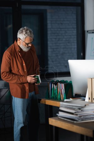 Photo for Grey haired businessman holding notebook while standing near documents and computer monitor in office - Royalty Free Image