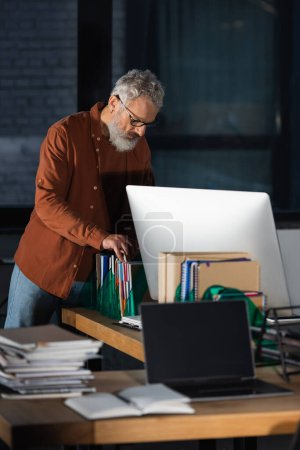 Téléchargez les photos : Homme d'affaires mature travaillant avec des papiers près des ordinateurs dans le bureau sombre - en image libre de droit