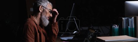 Foto de Vista lateral del hombre de negocios cansado y reflexivo tocando la frente mientras trabaja en el ordenador portátil en la oficina oscura, pancarta - Imagen libre de derechos