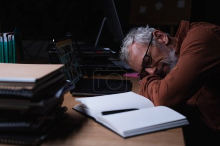 mature exhausted businessman sleeping near notebook and laptop in office at night