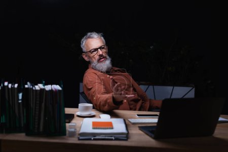 bärtiger Geschäftsmann mit Brille zeigt mit der Hand, während er während eines Videochats auf dem Laptop im dunklen Büro spricht