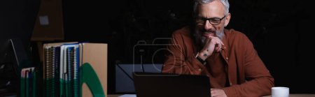 Photo for Mature and bearded businessman in eyeglasses smiling during video call in office at night, banner - Royalty Free Image