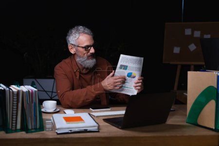 Photo for Middle aged businessman holding paper with graphs during video chat on laptop in office at night - Royalty Free Image