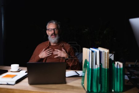 zufriedener Geschäftsmann berührt Brust und schaut auf Laptop neben Kaffeetasse und Notizbücher im dunklen Büro