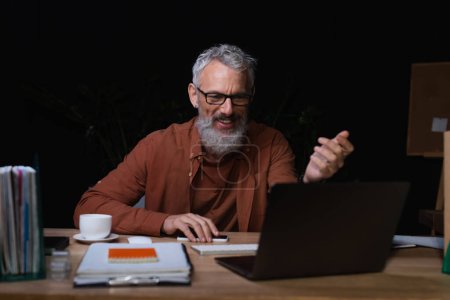 glücklicher grauhaariger Geschäftsmann gestikuliert bei Videochat auf Laptop neben Kaffeetasse im dunklen Büro