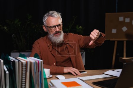 grey haired bearded businessman showing thumb down during video call on laptop in office at night
